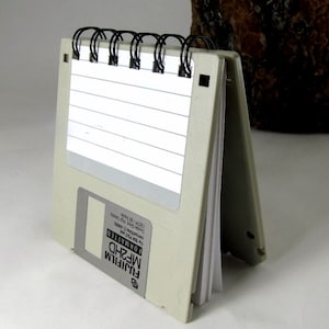 May include: A gray and white notebook with a spiral binding. The notebook is made from a repurposed floppy disk. The floppy disk is labeled "FUJIFILM MF2HD FORMATTED For IBM PS/2 and compatibles 1.44MB 135TPI Double Sided High Density".