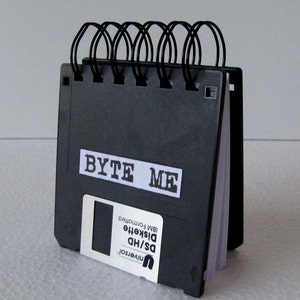 May include: A black notebook with a black and white label that says "BYTE ME". The notebook is made from recycled floppy disks and has a black spiral binding.