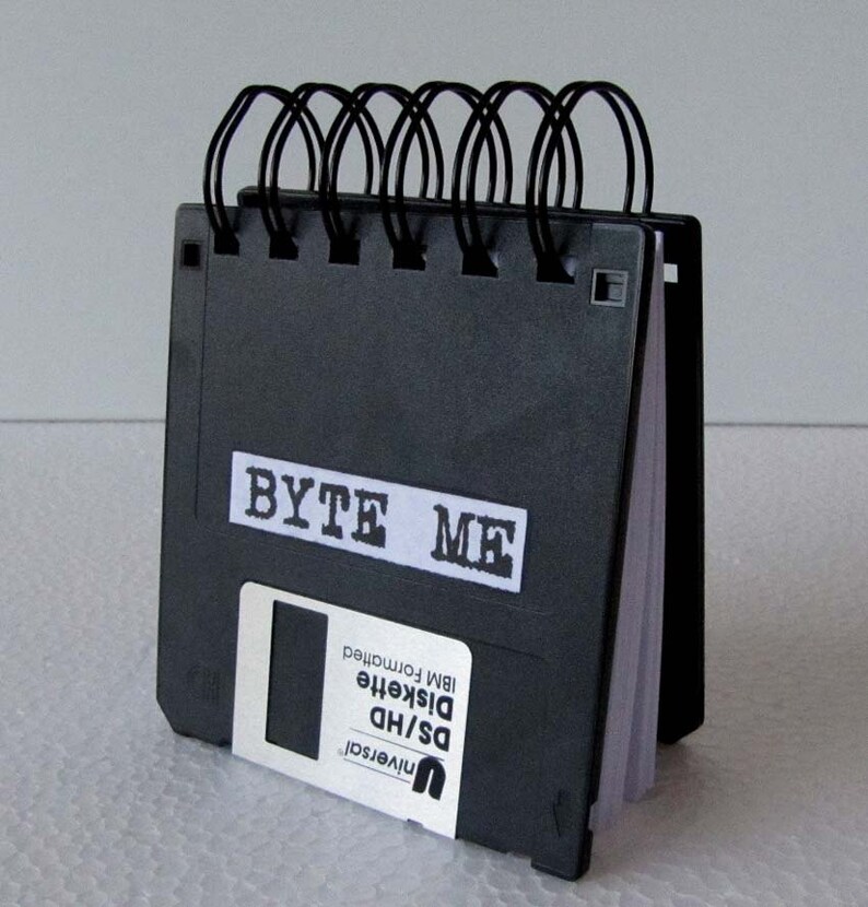 May include: A black notebook with a black and white label that says "BYTE ME". The notebook is made from recycled floppy disks and has a black spiral binding.