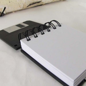 May include: A black spiral-bound notebook with white pages, resting on a black 8.89 cm floppy disk. The notebook's binding is black, and the pages are blank. A piece of birch bark is in the background.