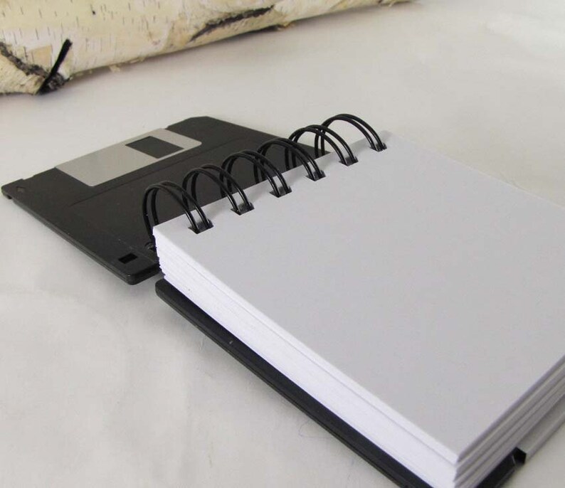 May include: A black spiral-bound notebook with white pages, resting on a black 8.89 cm floppy disk. The notebook's binding is black, and the pages are blank. A piece of birch bark is in the background.