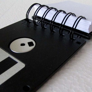 May include: A black floppy disk with a silver label and a black spiral notebook attached to it.