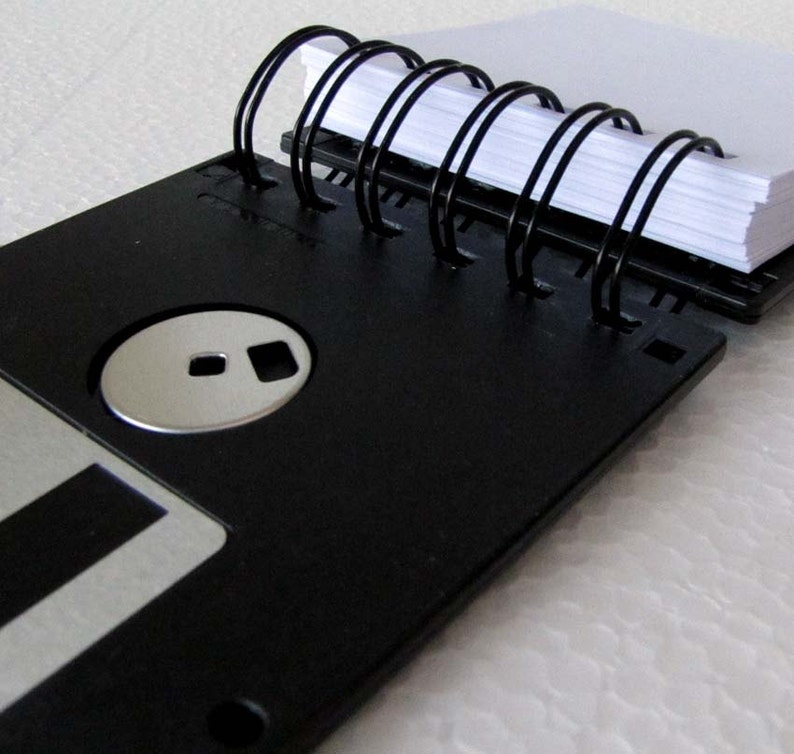 May include: A black floppy disk with a silver label and a black spiral notebook attached to it.