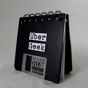 May include: A black notebook with a spiral binding. The cover is made from a recycled floppy disk. The notebook has the words "Uber Geek" printed on the front.