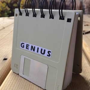 Peut inclure: Un petit carnet à spirales gris avec un design de disquette. Le carnet porte l'inscription "GENIUS" sur la couverture.