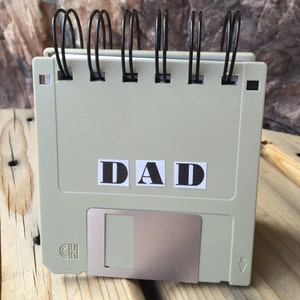 May include: A grey floppy disk with a metal insert and a black spring binder. The disk has the word "DAD" printed on it in black letters.