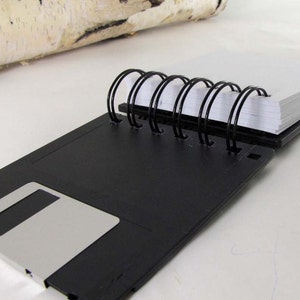 May include: A black notebook made from a repurposed floppy disk, featuring a silver rectangular detail. It has a spiral binding and contains white paper pages. A piece of birch bark is visible in the background.