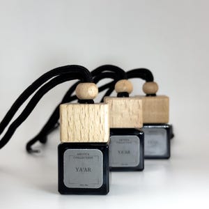 May include: Three black car air fresheners with wooden tops and black cords. Each has a silver label with the text "ARUCCA COLLECTION" and "YA'AR". The fresheners are arranged in a row, with the front one in focus.