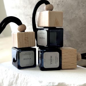 May include: A stack of four dark blue glass car air fresheners with wooden tops and black cords. Each square bottle has a label that reads "ARUCCA COLLECTION HEAYLA". The fresheners are displayed on a white, textured surface.