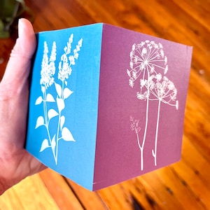 May include: A folded card with a blue and purple background. The front of the card features a white silhouette of a flower on the blue side and a white silhouette of a different flower on the purple side.