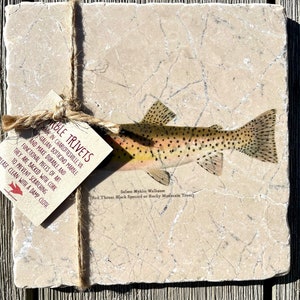 May include: A set of two marble trivets featuring a brown trout with black spots on a beige marble background. The trivets are made in Charlottesville, Virginia, and are backed with cork to prevent scratching. The text "Able Trivets" and "Salmo Mykiss Walbaum (Red Throat Black Spotted or Rocky Mountain Trout)" is printed on the trivets.