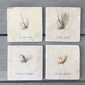 May include: Four square ceramic coasters with images of different fishing flies. The coasters are beige with a light brown marbled pattern. The flies are depicted in detail with realistic colors and textures. The coasters are labeled with the names of the flies: Stone Fly, Grey Drake, Brown Palmer, and Red Spinner.