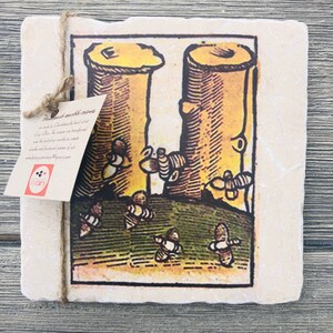 May include: A square, beige marble coaster with a vintage illustration of two tree trunks with bees flying around them. The text "Destande marble trivet" is printed on a white tag attached to the coaster.