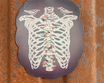 May include: A sticker with a white ribcage design on a dark purple background. The ribcage is surrounded by a vine with pink flowers and butterflies. Two small white birds are perched on the top of the ribcage.