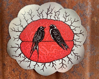 May include: A silver sticker with a red and white design of two birds in a nest. The birds are black with white accents and are facing each other. The nest is surrounded by branches.