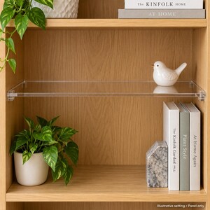 May include: A clear acrylic shelf panel mounted horizontally within a light wood bookcase. The transparent rectangular shelf features small, clear mounting brackets on each side, providing a floating appearance for displaying decorative items.