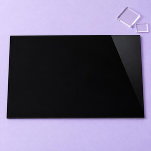 May include: A large, rectangular, solid black acrylic sheet with a smooth, reflective surface. Positioned near the top right corner are two smaller, clear, square acrylic pieces, one slightly larger than the other, resting on a flat surface.
