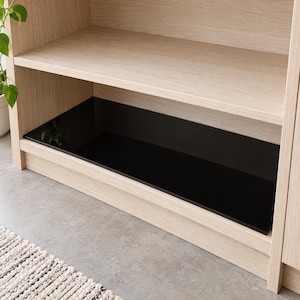 May include: A light-colored wood shelving unit featuring a smooth, light-toned wood grain finish on the frame and shelves, with a solid, reflective black rectangular panel installed on the bottom shelf surface.