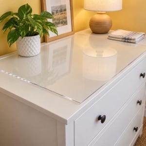 May include: A clear rectangular acrylic protective cover placed on top of a white wooden dresser with three drawers. The dresser features dark round knobs and a smooth surface, providing a transparent layer of protection for the furniture top.