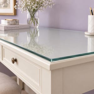 May include: A white wooden desk featuring a clear glass top, a single drawer with a round brass-colored knob, and a light-colored upholstered stool. The desk surface holds a white marble pen holder and a book titled The Inspired Home.