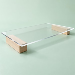 May include: A rectangular clear acrylic tray featuring two light-toned wooden block supports at opposite ends. The transparent surface is smooth and flat, resting securely on the two solid wooden bases, creating a raised platform design.