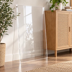 May include: A large, rectangular, clear acrylic sheet standing upright on a floor. The transparent panel features smooth, polished edges and a flat, reflective surface that captures light and shadows from the surrounding environment.
