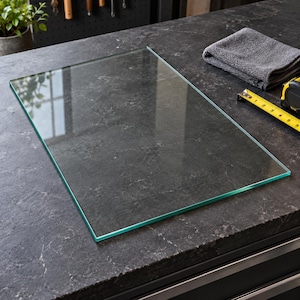 May include: A rectangular, clear glass cutting board with smooth, polished edges. The transparent surface is flat and features a clean, minimalist design, allowing the underlying dark, textured countertop to be clearly visible through the glass.