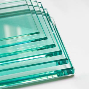 May include: A stack of multiple rectangular clear glass sheets with a distinct light green tint along the edges. Each sheet features smooth, polished surfaces and straight, sharp corners, creating a layered, transparent appearance.