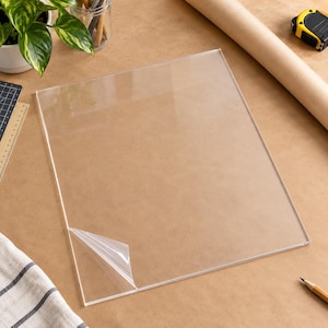 May include: A rectangular clear acrylic sheet with a smooth, transparent surface. One corner of the protective film is partially peeled back, revealing the glossy material underneath. The sheet is flat and rigid, featuring sharp, clean edges.