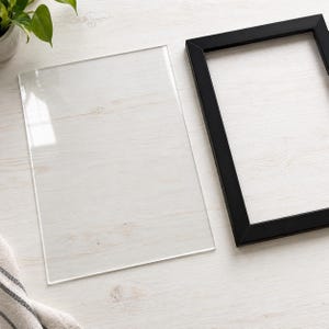 May include: A rectangular clear acrylic or glass sheet positioned next to an empty black rectangular picture frame. Both items are rectangular in shape and feature clean, sharp edges, presented as separate components for framing.