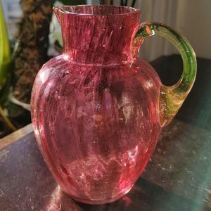 May include: A pink glass jug with a green handle. The jug has a textured surface and a rounded body.