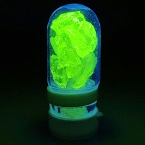 May include: A luminous green mineral sample contained within a clear, cylindrical vial with a blue cap. The base of the vial is light green. The mineral exhibits a rough, crystalline form and glows brightly against a dark backdrop.