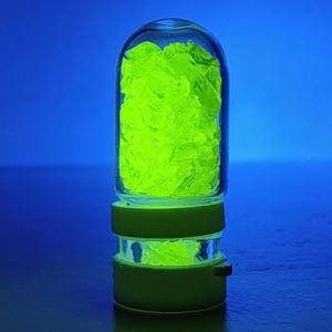 May include: A glass jar filled with bright green crystals. The jar has a clear glass body and a green plastic lid.