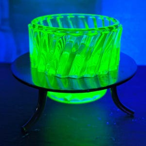 May include: A vibrant green glass dish with a spiral pattern is displayed on a small black metal stand with three curved legs. The dish and stand are lit with a bright green light, creating a striking visual effect.