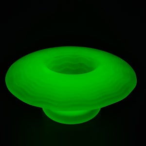 May include: A glowing green glass bowl with a rounded, saucer-like shape. The bowl has a smooth, frosted finish and a central opening.