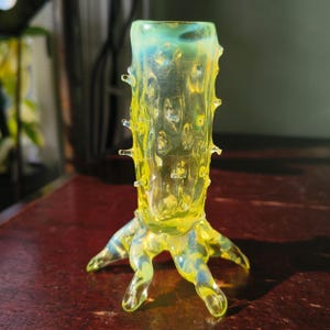 May include: A distinctive, hand-blown glass vase in yellow and blue hues. The vase features a textured, tree-like design with small protrusions and a root-like base. The translucent glass allows light to pass through, creating a luminous effect.