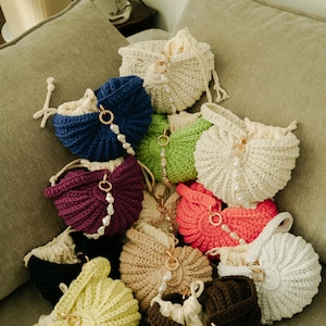 May include: A collection of handmade crochet drawstring bags in various colors, including blue, green, pink, and cream. Each bag features a pearl accent and a gold-colored ring. The bags are arranged on a neutral-colored surface.