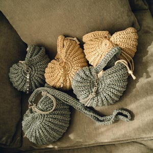 May include: An overhead shot displays several handmade crochet bags in shades of gray and tan. Some bags have beaded accents and drawstrings. One gray bag has a long strap. The bags are arranged on a soft, olive-green surface.