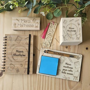 Peut inclure: Ensemble de cadeaux en bois pour remercier une maîtresse. Comprend un bloc-notes, un tampon, un marque-page, un stylo et un bloc avec les mots "Merci Maîtresse" et des illustrations scolaires. Un bloc-notes adhésif bleu est également inclus.