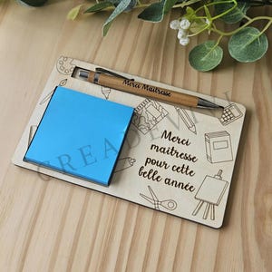 Può includere: Un organizer da scrivania in legno con un blocco note blu e una penna in bambù. La penna è incisa con "Merci Maitresse". L'organizer presenta illustrazioni di materiale scolastico e il testo "Merci maitresse pour cette belle année".