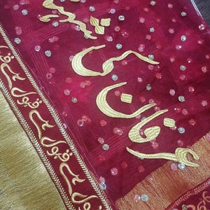 May include: A red fabric shawl with gold embroidery and sequins. The embroidery features Arabic calligraphy.