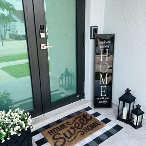 May include: A black and white checkered welcome mat with the words "Home Sweet Home" in brown script. There is a black sign with white lettering that reads "It's for me and my HOME we will serve the Lord" and two black lanterns with candles.
