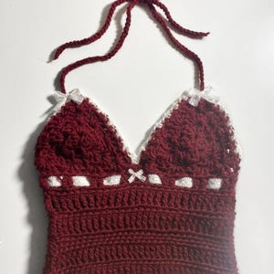 Puede incluir: Un top halter de color granate tejido a crochet con ribetes y lazos de crochet blanco. El top tiene un escote en V y tirantes ajustables.