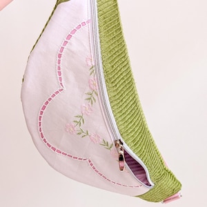 May include: A light pink and green corduroy fanny pack with a floral embroidered design. The bag has a pink strap and zipper, and a silver zipper. The bag is held by a hand.