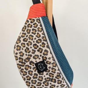Könnte beinhalten: Gürteltasche aus Cord mit Leopardenmuster, orangefarbenen und tealfarbenen Akzenten. Die Tasche hat einen silbernen Reißverschluss und einen kleinen schwarzen Aufnäher mit einer Katzenillustration. Die Tasche hängt an einer Holzstange.