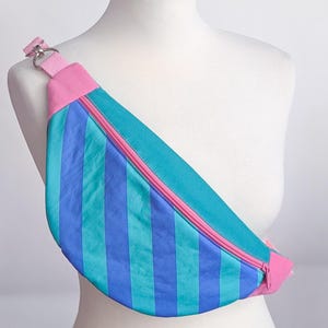 May include: A colorful fanny pack with vertical stripes in shades of blue and teal, accented with a pink zipper and strap. The bag is displayed on a white mannequin, showcasing its design and shape.