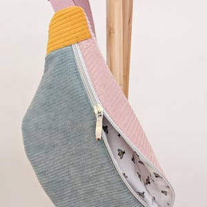 Könnte beinhalten: Eine hellblaue, rosafarbene und gelbe Cord-Bauchtasche mit Reißverschluss und einem weißen Futter mit Bienenmuster.