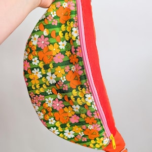 May include: A colorful fanny pack with a floral pattern. The bag features a green background with orange, pink, yellow, and white flowers. It has an orange strap and trim, a pink zipper, and a yellow zipper pull.