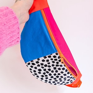 Peut inclure: Un sac banane coloré au design vif. Le sac présente une combinaison de couleurs vives, notamment orange, rose et bleu, avec une section en tissu à pois noirs et blancs. Il possède une fermeture éclair et une sangle orange.