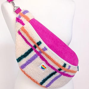 May include: A fanny pack made of cream-colored sherpa fleece featuring a plaid pattern in orange, purple, and dark green. The bag has a bright pink corduroy top panel, a matching pink adjustable strap, and a small white label with a rainbow heart.